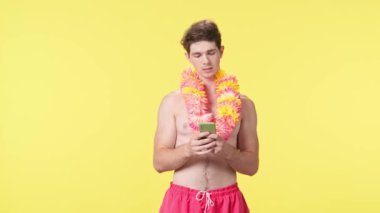 Genç adam akıllı telefonuyla mesaj atıyor. Pembe şortlu çıplak gövdeli erkek manken ve sarı arka planda Hawai lei pozu. Seyahat, yaz tatili, plaj eğlencesi konsepti.