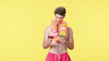 Genç erkek akıllı telefon ve banka kartıyla online alışveriş yapıyor. Pembe şortlu çıplak gövdeli erkek manken ve sarı arka planda Hawai lei pozu. Seyahat, yaz tatili, plaj tatili.