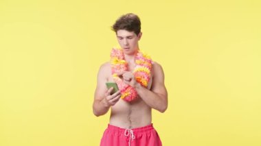 Genç bir erkek model bir akıllı telefona bakar ve kazanma ya da iyi haber karşısında sevinir. Pembe şortlu çıplak gövdeli erkek manken ve sarı arka planda Hawaii lei pozları..