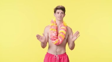 Genç bir adam kameraya bakar, bir şey söyler ve elleriyle hareket eder. Pembe şortlu çıplak gövdeli erkek manken ve sarı arka planda Hawaii lei pozları..