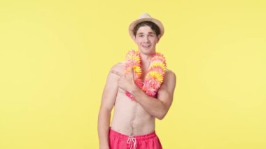 Genç erkek model, parmağını işyerine doğrultuyor ve baş parmak hareketi yapıyor. Pembe şortlu çıplak gövdeli erkek model ve sarı arka planda Hawaii lei pozları..