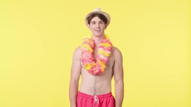 Genç bir erkek kameraya büyük bir zevkle bakıyor. Pembe şortlu çıplak gövdeli erkek manken ve sarı arka planda Hawai lei pozu. Seyahat, yaz tatili, plaj eğlencesi konsepti.