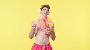 Genç adam akıllı telefon kullanarak kulaklıkla müzik dinliyor. Pembe şortlu çıplak gövdeli erkek manken ve sarı arka planda Hawai lei pozu. Seyahat, yaz tatili.