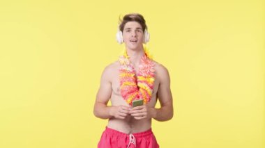 Genç adam akıllı telefon kullanarak kulaklıkla müzik dinliyor. Pembe şortlu çıplak gövdeli erkek manken ve sarı arka planda Hawai lei pozu. Seyahat, yaz tatili.