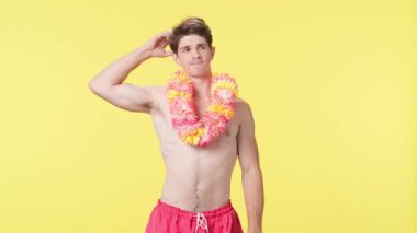 Genç adam bir şey düşünüyor ve hiçbir fikri olmadığı için hayal kırıklığına uğruyor. Pembe şortlu çıplak gövdeli erkek manken ve sarı arka planda Hawaii lei pozu. Seyahat, yaz tatili.