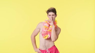 Genç adam kameraya bakarak poz veriyor. Pembe şortlu çıplak gövdeli erkek manken ve sarı arka planda Hawai lei pozu. Seyahat, yaz tatili, sahil tatili konsepti.