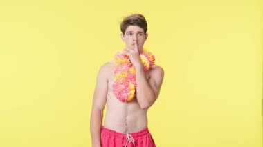 Genç adam bir şey düşünüyor ve sonra biletleri tatil için iyi bir fikir olarak gösteriyor. Pembe şortlu çıplak bir adam ve sarı arka planda Hawaii lei pozu. Seyahat, yaz tatili.