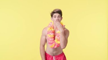 Genç bir adam hava öpücüğü gönderir ve parmaklarıyla bir kalp sembolü gösterir. Pembe şortlu çıplak bir adam ve sarı arka planda Hawaii lei pozu veren bir adam. Seyahat, yaz tatili.