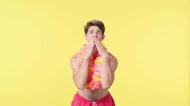 Genç bir adam hava öpücüğü gönderir ve parmaklarıyla bir kalp sembolü gösterir. Pembe şortlu çıplak bir adam ve sarı arka planda Hawaii lei pozu veren bir adam. Seyahat, yaz tatili.