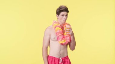 Kokteyl içen genç bir adam. Tadı ve tazeliği onu çok mutlu ediyor. Pembe şortlu çıplak bir adam ve sarı arka planda Hawaii lei pozu. Seyahat, yaz tatili.