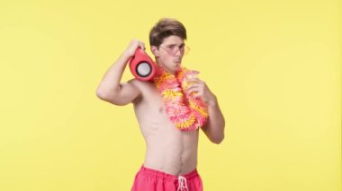 Genç bir adam kokteyl içiyor ve taşınabilir bir ses hoparlörünün müziğiyle dans ediyor. Pembe şortlu çıplak bir adam ve sarı arka planda Hawaii lei pozu veren bir adam. Seyahat, yaz tatili.