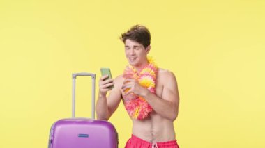 Genç adam akıllı telefonuyla mesaj atıyor ve kokteyl içiyor. Pembe şortlu tişörtsüz bir adam ve sarı arka planda poz veren Hawaii zambağı. Seyahat, yaz tatili.
