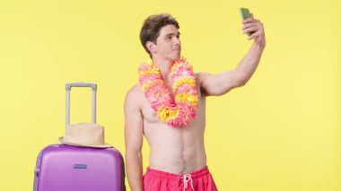 Genç adam akıllı telefonuyla poz veriyor ve selfie çekiyor. Pembe şortlu tişörtsüz bir adam ve sarı arka planda poz veren Hawaii zambağı. Seyahat, yaz tatili.