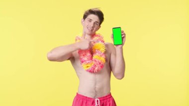 Genç adam yeşil ekranlı akıllı telefon ekranını işaret ediyor. Sarı arka planda pembe şortlu çıplak bir adam poz veriyor. Seyahat, yaz tatili.