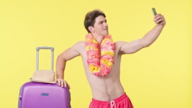 Genç adam akıllı telefonuyla poz veriyor ve selfie çekiyor. Pembe şortlu tişörtsüz bir adam ve sarı arka planda poz veren Hawaii zambağı. Seyahat, yaz tatili.