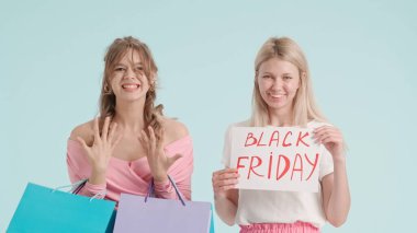 Stüdyoda poz veren iki genç kadın. Genç modaya uygun kadın modeller, mavi arka planda izole edilmiş alışveriş çantaları ve satış tabelasıyla poz veriyorlar. Moda ve gençlik güzellik konsepti.