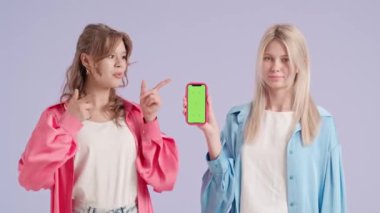 Stüdyoda poz veren iki genç kadın. Mor arka planda izole edilmiş akıllı telefon kroma anahtar yeşil ekran gösteren genç ve şık kadın modeller. Moda ve gençlik güzellik konsepti.