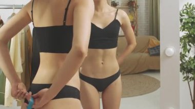Evde aynanın önünde poz veren genç bir kadın model. Genç milenyum kadını yatak odasında aynaya bakıyor, bel ölçüsünü ölçüyor, mutlu bir yüz. Kişisel bakım sağlıklı güzellik konsepti.