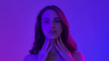 Çekici kadın model kameraya güzellik tedavisi yapıyor. Kameraya poz veren genç bir kadın pürüzsüz tenine dokunuyor ve gülümsüyor, mavi neon arka planda izole edilmiş. Doğal güzellik ve cilt bakımı kavramı