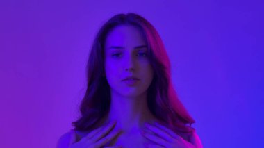 Çekici kadın model kameraya güzellik tedavisi yapıyor. Kameraya poz veren genç bir kadın pürüzsüz tenine dokunuyor ve gülümsüyor, mavi neon arka planda izole edilmiş. Doğal güzellik ve cilt bakımı kavramı