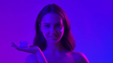 Çekici kadın model kameraya güzellik tedavisi yapıyor. Kameraya poz veren genç kadın elinde kavanozla cildine kozmetik ürünler uyguluyor, mavi neon arka planda izole edilmiş. Doğal güzellik cilt bakımı kavramı