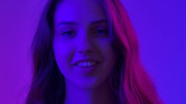 Genç bir kadın kameranın önünde poz veriyor ve tatlı tatlı gülümsüyor, mavi neon arka planda izole bir şekilde. Doğal güzellik ve cilt bakımı kavramı.