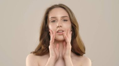 Çekici kadın model kameraya güzellik tedavisi yapıyor. Kameraya poz veren genç bir kadın beyaz arka planda izole edilmiş pürüzsüz tenine dokunuyor. Doğal güzellik cilt bakımı kavramı