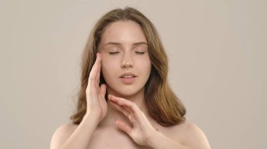 Çekici kadın model kameraya güzellik tedavisi yapıyor. Kameraya poz veren genç bir kadın beyaz arka planda izole edilmiş pürüzsüz tenine dokunuyor. Doğal güzellik cilt bakımı kavramı