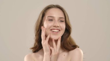 Çekici kadın model kameraya güzellik tedavisi yapıyor. Kameraya poz veren genç bir kadın beyaz arka planda izole edilmiş pürüzsüz tenine dokunuyor. Doğal güzellik cilt bakımı kavramı