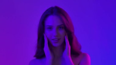 Çekici kadın model kameraya güzellik tedavisi yapıyor. Kameraya poz veren genç bir kadın pürüzsüz tenine dokunuyor ve gülümsüyor, mavi neon arka planda izole edilmiş. Doğal güzellik ve cilt bakımı kavramı