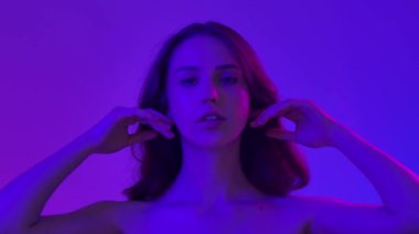 Çekici kadın model kameraya güzellik tedavisi yapıyor. Kameraya poz veren genç bir kadın pürüzsüz tenine dokunuyor ve gülümsüyor, mavi neon arka planda izole edilmiş. Doğal güzellik ve cilt bakımı kavramı