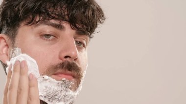 Gömleksiz, yakışıklı, sakallı erkek model sabah güzellik gösterisi yapıyor. Tıraş köpüğü süren genç bir adam beyaz arka planda izole edilmiş kameraya bakıyor. Doğal güzellik cilt bakımı kavramı.