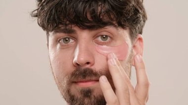 Gömleksiz, yakışıklı, sakallı erkek model sabah güzellik gösterisi yapıyor. Gözlerinin altına göz bandı süren genç bir adam beyaz arka planda izole bir şekilde kameraya bakıyor. Doğal güzellik cilt bakımı kavramı.