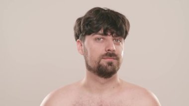 Gömleksiz, yakışıklı, sakallı erkek model sabah güzellik gösterisi yapıyor. Genç bir adam tıraş kremi sürerken beyaz arka planda izole edilmiş kameraya bakar. Doğal güzellik cilt bakımı kavramı