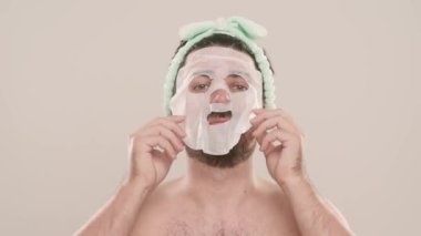 Gömleksiz, yakışıklı, sakallı erkek model sabah güzellik gösterisi yapıyor. Genç bir adam beyaz arka planda izole edilmiş kameraya bakarak yüz maskesi takıyor. Doğal güzellik cilt bakımı kavramı.