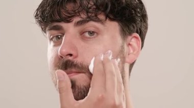 Gömleksiz, yakışıklı, sakallı erkek model sabah güzellik gösterisi yapıyor. Tıraş köpüğü süren genç bir adam beyaz arka planda izole edilmiş kameraya bakıyor. Doğal güzellik cilt bakımı kavramı.