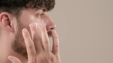 Gömleksiz, yakışıklı, sakallı erkek model sabah güzellik gösterisi yapıyor. Genç bir adam tıraş kremi sürerken beyaz arka planda izole edilmiş kameraya bakar. Yan görünüm.