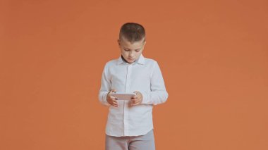 Akıllı telefondan oyun oynayan genç bir çocuk var. Stüdyoda turuncu arka planda poz veren beyaz gömlekli öğrenci. Ortaokul ve eğitim konsepti.