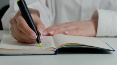 Masadaki kadını yakın plan çekimde elinde sarı kalemle defter, okuma ve önemli kısımları işaretlerken çek..