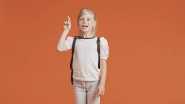 Genç kız bir şey düşünüyor ve bir jestle bir fikri olduğunu gösteriyor. Stüdyoda turuncu arka planda poz veren kız öğrenci. Ortaokul ve eğitim kavramı.