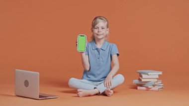 Elinde yeşil ekran krom anahtarı olan akıllı telefonu tutan ve baş parmağını gösteren genç kız. Stüdyoda turuncu arka planda poz veren kız öğrenci. İlkokul ve eğitim konsepti.