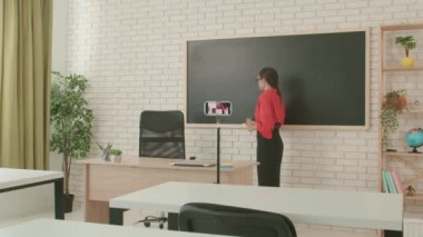 Okulda öğretmen video konferansında. Boş sınıftaki kadın öğretmen, akıllı telefon kullanarak canlı video dersi konferansını canlı yayınlıyor. Uzak bir uzak modern eğitim.
