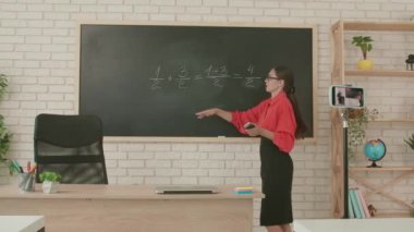 Okulda öğretmen video konferansında. Boş sınıftaki kadın öğretmen matematik örneğini açıklamak için akıllı telefon kullanarak canlı video dersi konferansı yapıyor. Uzak bir uzak modern eğitim.