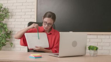 Okulda öğretmen video konferansında. Boş sınıftaki kadın öğretmen dizüstü bilgisayarla canlı video dersi konferansında kağıt üzerinde materyal gösteriyor. Uzak bir uzak modern eğitim.