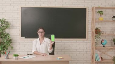 Okul öğretmeni sınıfta gençlerle konuşuyor. Sınıfta oturan kadın kolej öğretmeni elinde krom anahtar yeşil ekran arka planlı akıllı telefonuyla eğitim konseptini gösteriyor..