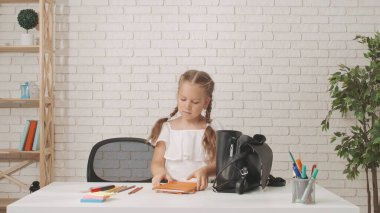 Küçük kız okula ya da anaokuluna gidiyor. Evdeki oturma odasındaki masada oturan tatlı kız, okula hazırlanan çocuk, sırt çantasına malzeme ve defterler koyuyor..