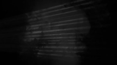 Bir adam yatay ışık şeritleriyle kısmen aydınlanarak dramatik gölgelerle yüzleşir. Karanlıktan ve dumandan bir adam silueti belirir, kameraya bakar ve şeytanca gülümser..