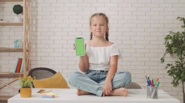 Küçük kız okula ya da anaokuluna gidiyor. Küçük kız oturma odasında masada oturuyor, elinde krom anahtarlı akıllı telefon, yeşil ekran arka plan modeli ve gülümsüyor..