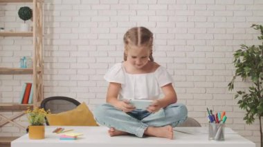 Küçük kız okula ya da anaokuluna gidiyor. Küçük kız oturma odasında masada oturuyor, elinde akıllı bir telefon, internette video oyunu oynuyor, eğleniyor..