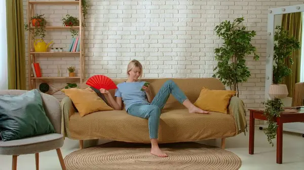 Stock image Young female spending time at home. Young millennial woman sitting on the sofa in the living room waving fan coping with heat and texting on smartphone. Stuffiness everyday life concept.